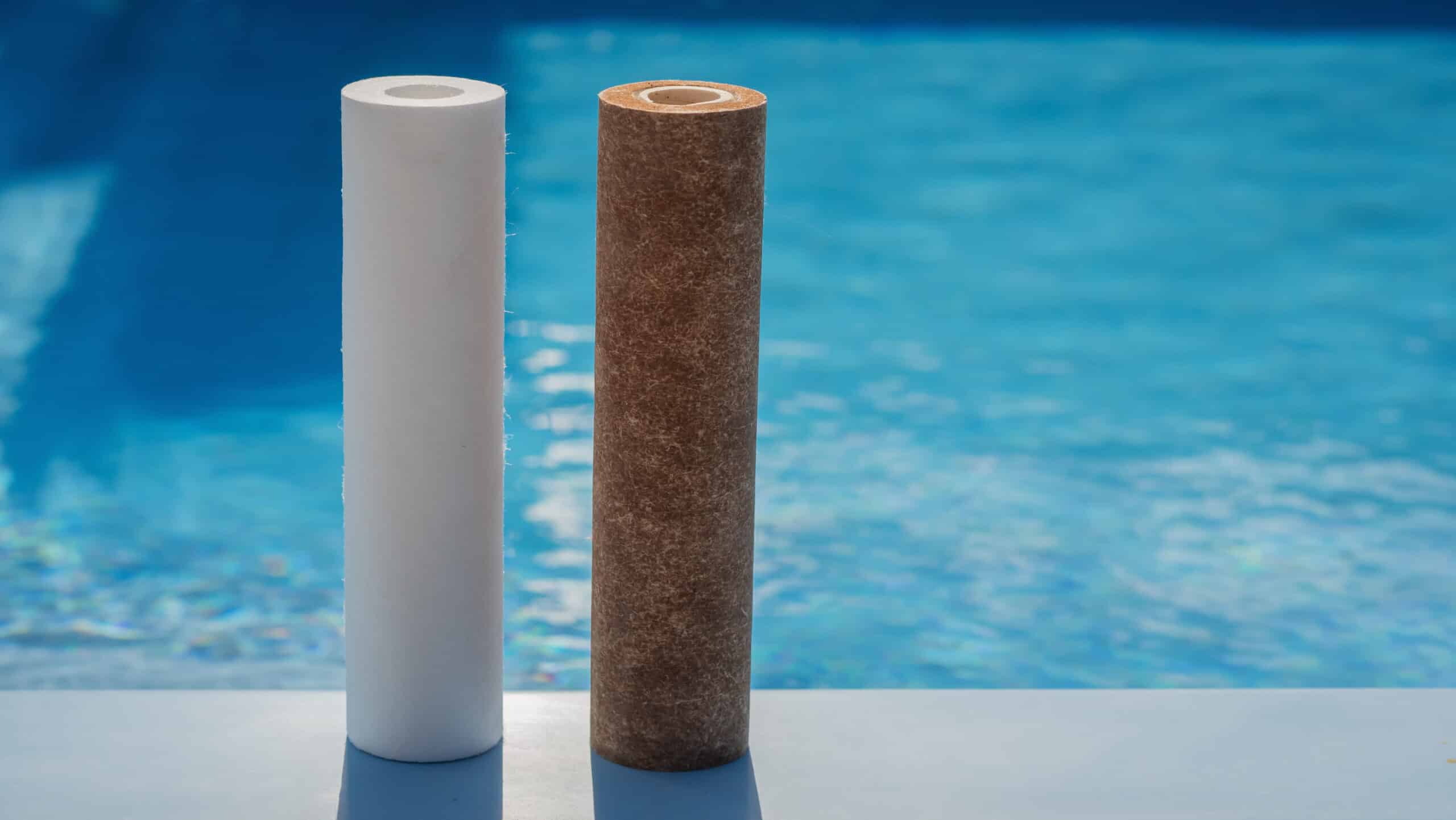 Two water filter cartridges are positioned upright by the edge of a swimming pool. One cartridge is clean and white, while the other is dirty and brown, showcasing the contrast between a new and used filter. The blue water in the pool background emphasizes the significance of clean filters in maintaining optimal water quality.